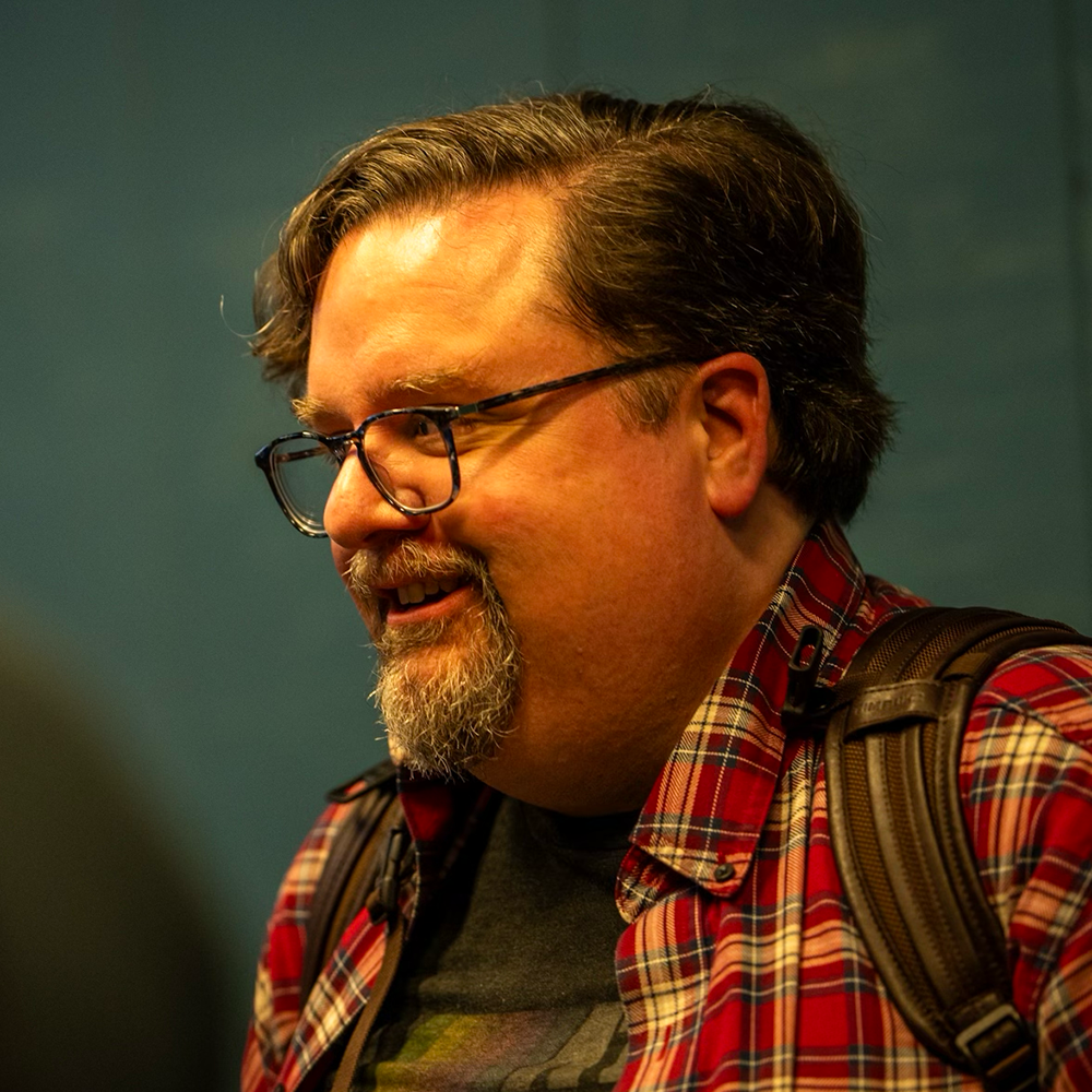 Chris Higgins wears a re checkered shirt and grins at someone off-camera. He has brown wavy hair and wears glasses, along with a brown and gray goatee.