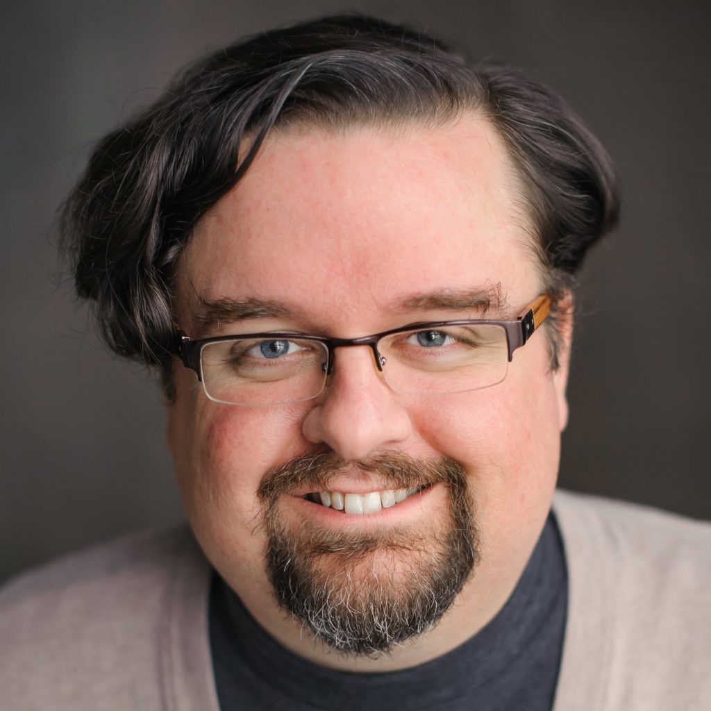 Portrait of Chris Higgins. He has a goatee, wears glasses, and smiles at the camera. He's wearing a turtleneck and a cardigan.